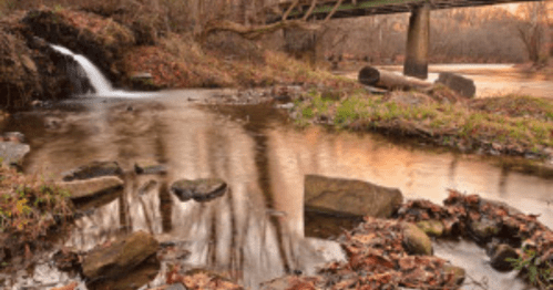 A serene landscape featuring a small waterfall, a calm stream, and a bridge in a wooded area during sunset.