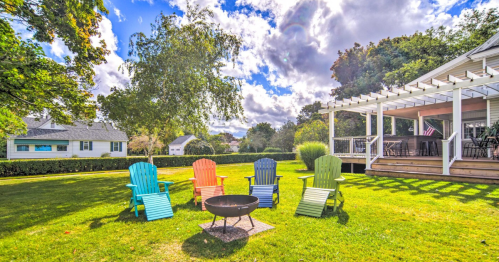 Colorful Adirondack chairs surround a fire pit in a sunny backyard with a deck and lush greenery.