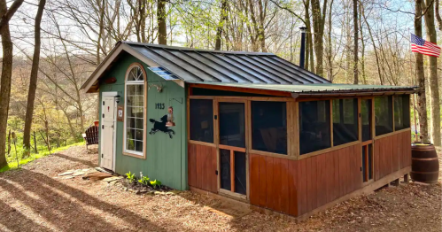 A small, charming cabin with a green and brown exterior, surrounded by trees and featuring a screened porch.