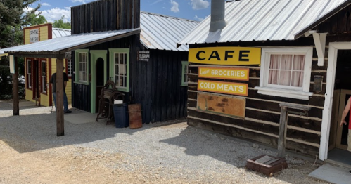 A rustic storefront with a café sign, featuring wooden buildings and a gravel path under a blue sky.