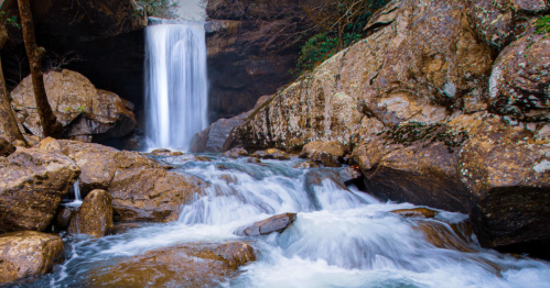 A serene waterfall cascades over rocks, surrounded by lush greenery and flowing water in a tranquil natural setting.