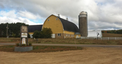 A yellow barn with a silo, surrounded by fields and trees, near a dirt road and a sign for a local business.