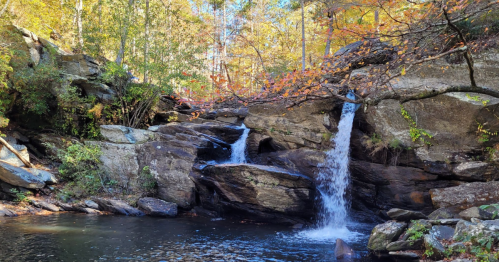 A serene waterfall cascading over rocks, surrounded by autumn foliage and a calm pool of water below.