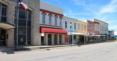 A quiet street lined with historic buildings and shops, featuring a Texas flag and clear blue skies.
