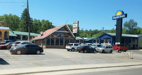 A Days Inn hotel with a café and parking lot filled with cars on a sunny day. Green trees in the background.