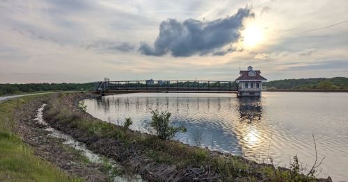 A serene lake scene featuring a bridge leading to a small building, with a cloudy sky and sun setting in the background.