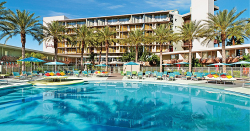 A sunny pool area with lounge chairs and palm trees, surrounded by a hotel building. Clear blue water reflects the sky.