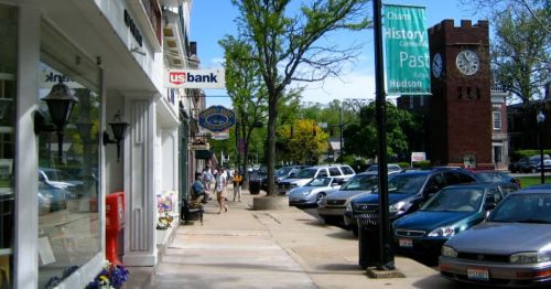 A sunny street scene with shops, parked cars, and a clock tower in the background. People walk along the sidewalk.