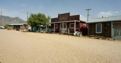 A dusty street scene featuring rustic buildings, including a red storefront and wooden houses, under a clear blue sky.