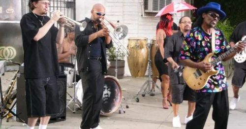 A lively band performs outdoors, featuring musicians with brass instruments, drums, and a guitarist in colorful attire.
