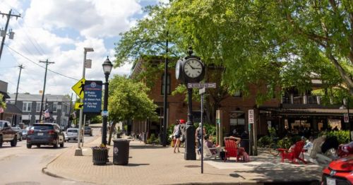 A lively street scene with a clock, trees, and people sitting outside a café on a sunny day.