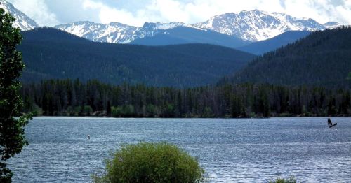 Scenic view of a lake surrounded by lush forests and snow-capped mountains under a partly cloudy sky.