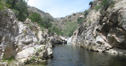 A serene river flows between rocky cliffs, surrounded by greenery and hills under a clear blue sky.