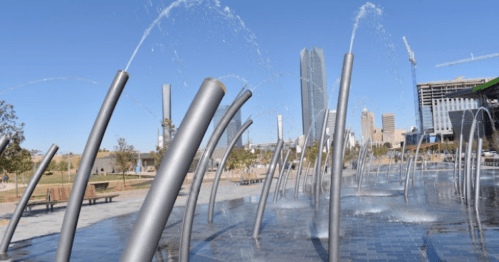 A modern fountain with arching water jets in a city park, surrounded by buildings and clear blue skies.