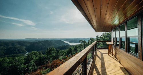 A wooden balcony overlooks a scenic view of mountains and a lake under a clear blue sky.