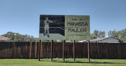 Sign reading "Home of Manassa Mauler Jack Dempsey" with an image of a boxer, set against a clear blue sky.