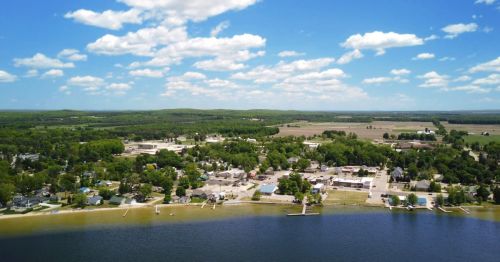 Aerial view of a small town by a lake, surrounded by green fields and hills under a blue sky with fluffy clouds.