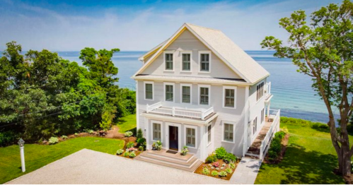 A large white house with a porch, surrounded by greenery, overlooking a calm body of water under a clear blue sky.