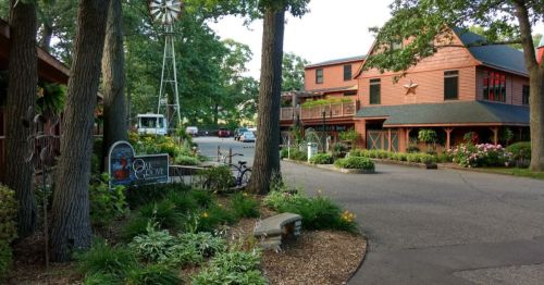 A scenic view of a rustic lodge surrounded by greenery, flowers, and a windmill in the background.
