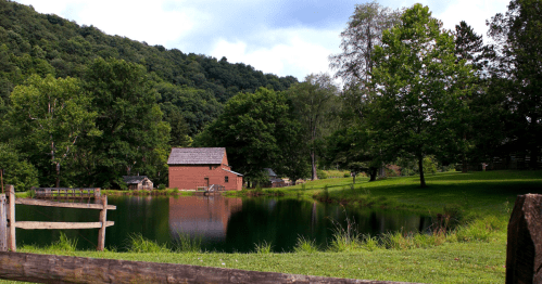 A serene landscape featuring a pond, a rustic cabin, and lush green trees under a cloudy sky.