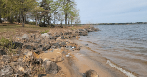 A serene lakeside view with rocky shorelines, sandy beach, and trees in the background under a cloudy sky.