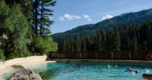 A serene outdoor pool surrounded by trees and mountains, with people swimming and enjoying the sunny day.