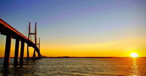 A bridge spans over water at sunset, with vibrant orange and blue hues in the sky reflecting on the surface.