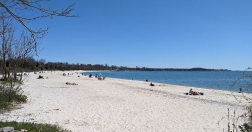 A sunny beach scene with people relaxing on the sand and calm water in the background under a clear blue sky.