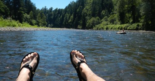Feet in sandals resting in a river, surrounded by lush green trees and a calm waterway.