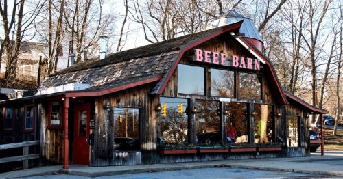 A rustic restaurant called "Beef Barn" with large windows, wooden exterior, and a sloped roof, surrounded by trees.