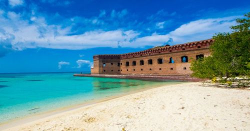 A historic brick fort on a sandy beach, surrounded by clear turquoise waters and a blue sky with scattered clouds.
