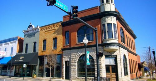 Historic brick buildings at a street corner, featuring a tall tower and storefronts under a clear blue sky.