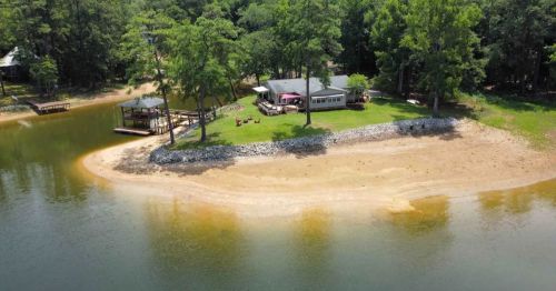 Aerial view of a lakeside property with a sandy shore, green lawn, and a house surrounded by trees.