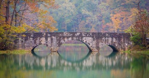 A stone bridge arches over a calm, reflective lake surrounded by autumn foliage.