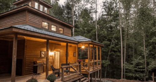 A wooden cabin in a forest, featuring a porch, outdoor seating, and large windows, surrounded by trees.