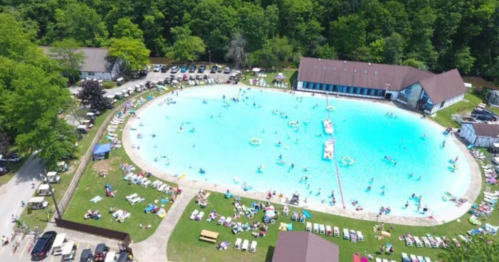 Aerial view of a large swimming pool surrounded by trees, with people swimming and lounging on the grass.