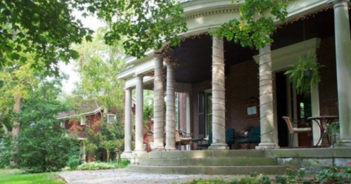 A charming porch with columns, surrounded by greenery and trees, leading to a cozy brick house in the background.