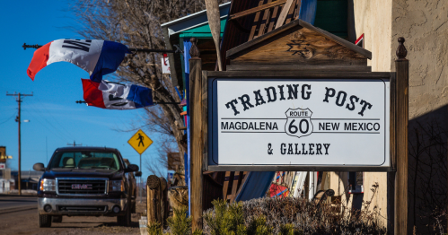 Sign for "Trading Post & Gallery" in Magdalena, New Mexico, with colorful flags and a pickup truck nearby.
