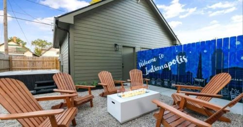 A cozy outdoor seating area with wooden chairs, a fire table, and a mural welcoming visitors to Indianapolis.