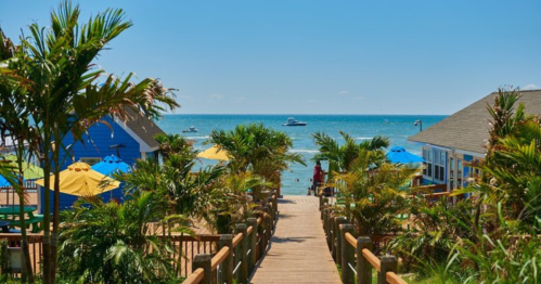 A wooden walkway leads to a beach with boats in the distance, surrounded by palm trees and colorful umbrellas.