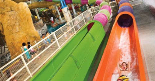 A child joyfully slides down a colorful water slide in a water park, surrounded by other attractions and visitors.