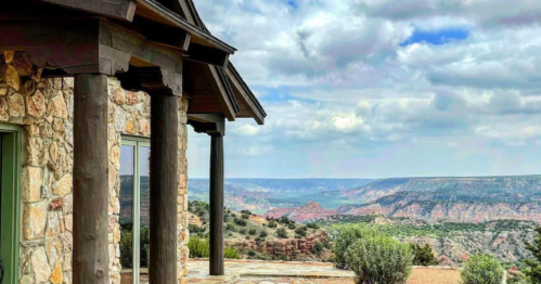 A stone house with wooden pillars overlooks a vast canyon under a cloudy sky.