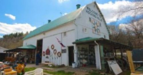 A white barn with a green roof, featuring a mural and various items displayed outside on a sunny day.
