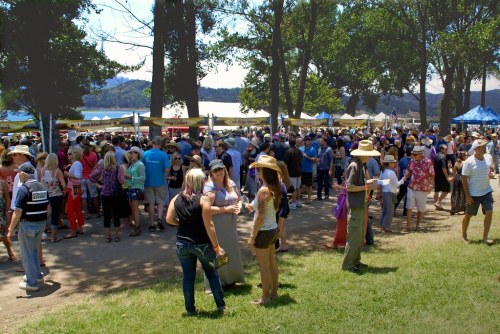 A crowded outdoor festival scene with people mingling under trees and tents, enjoying food and drinks.
