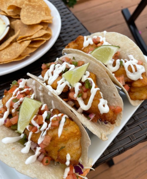 Three fish tacos topped with pico de gallo, lime wedges, and drizzled with creamy sauce, served with tortilla chips.