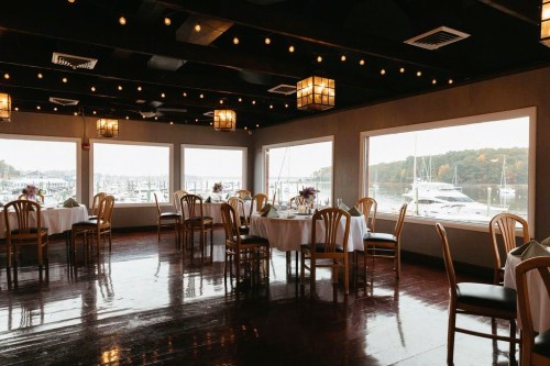 A cozy restaurant interior with tables set for dining, large windows overlooking a marina with boats.