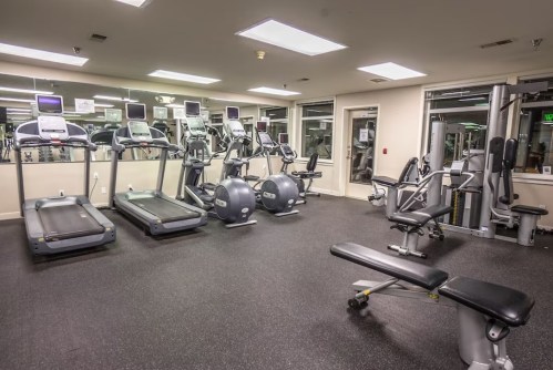 A modern gym with treadmills, stationary bikes, and weight benches on a rubber floor. Brightly lit with mirrors.