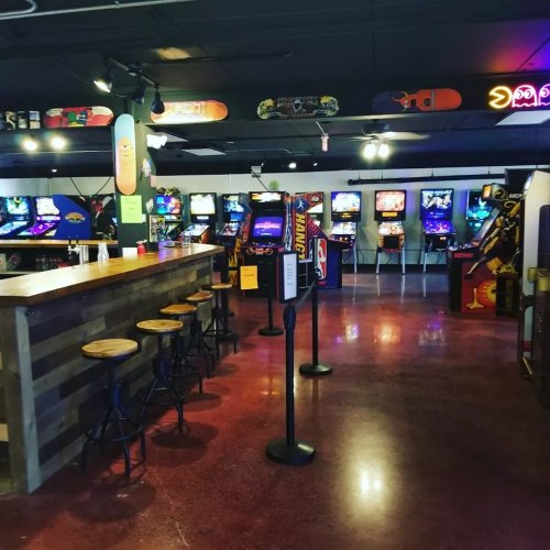 A vibrant arcade with various retro games, a wooden bar, and stools, featuring colorful neon lights and a lively atmosphere.