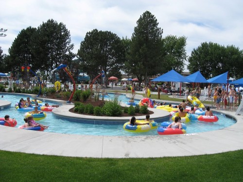 A busy water park scene with people in colorful inner tubes floating in a winding pool surrounded by trees and umbrellas.