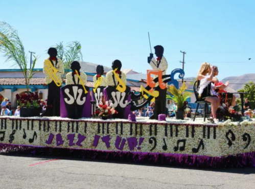 A colorful parade float featuring musicians playing instruments, surrounded by floral decorations and a cheering crowd.